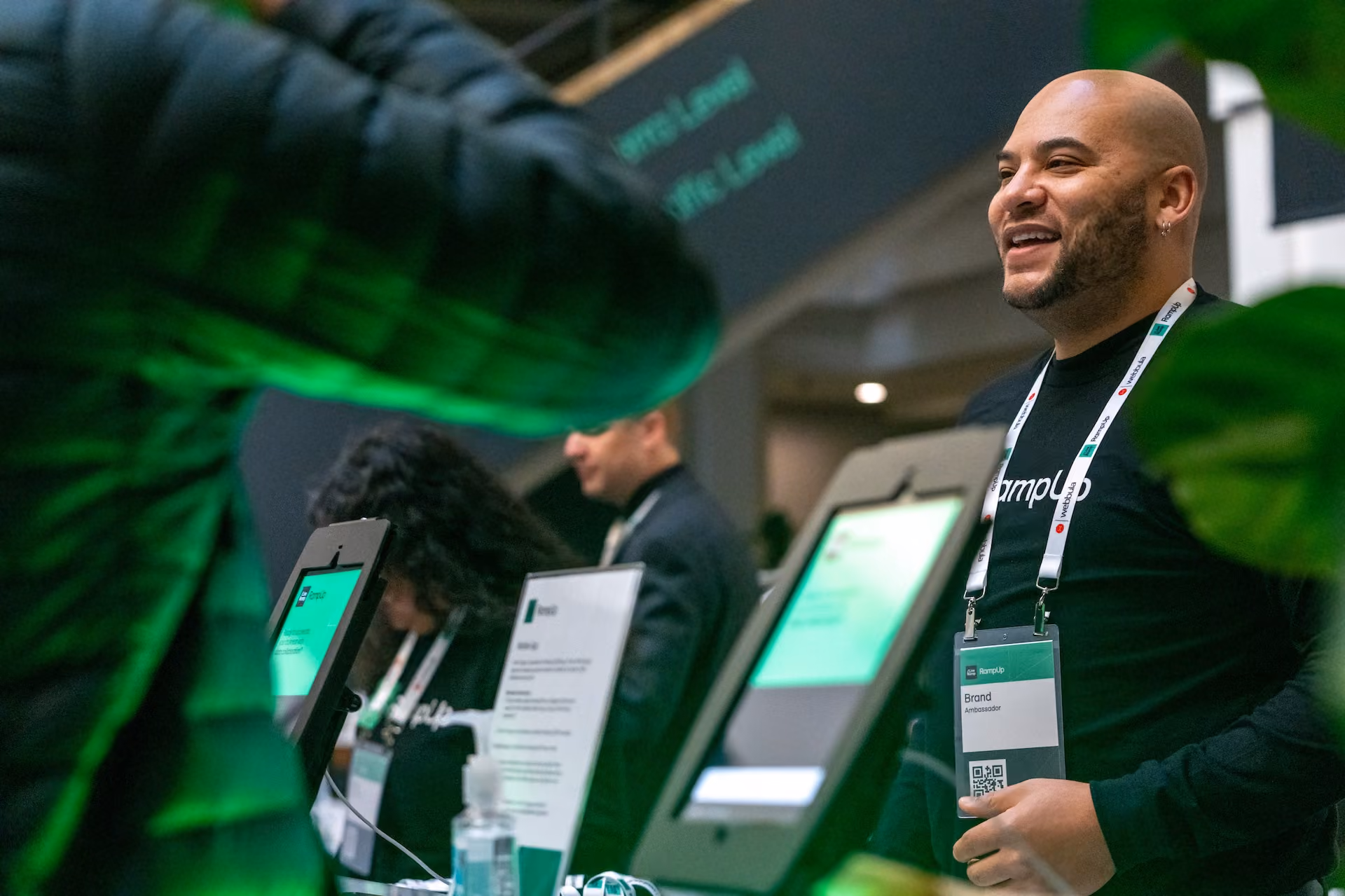 Smiling man wearing a black CampUp t-shirt and a lanyard with a badge labeled 'Brand Ambassador' standing at a booth with digital screens and other staff in black shirts in the background.
