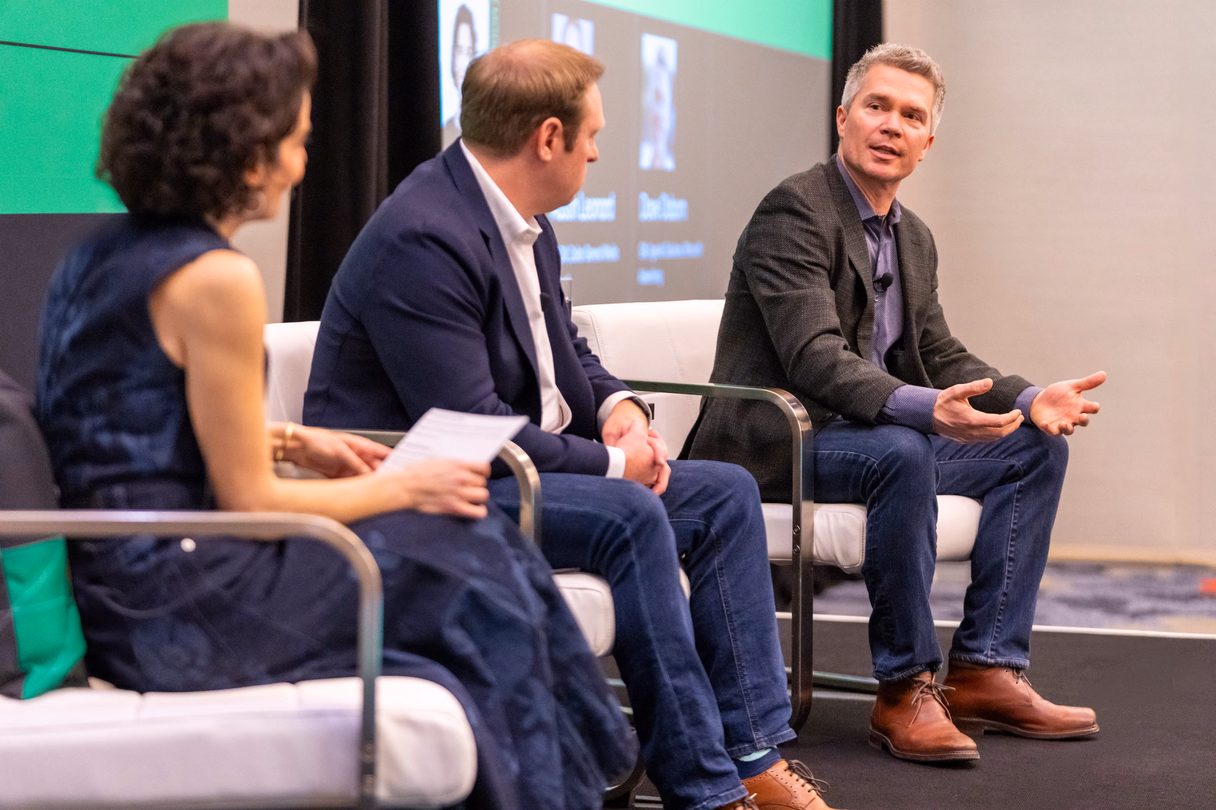 Three people seated on stage in a panel discussion, two men and one woman, with a screen showing blurred headshots and text in the background.