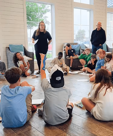 Group of children seated on floor in a bright room, some raising hands, with two adults standing.
