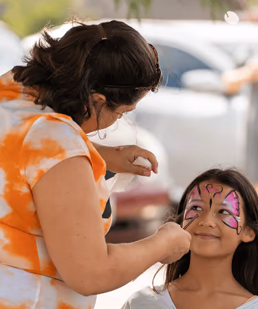 Woman painting colorful butterfly face art on smiling girl at outdoor event.