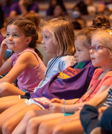 A group of young children sitting in a row, focused on an off-camera event or presentation.