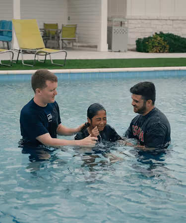 Two men in a pool gently holding a smiling girl between them underwater, with lawn chairs in background.