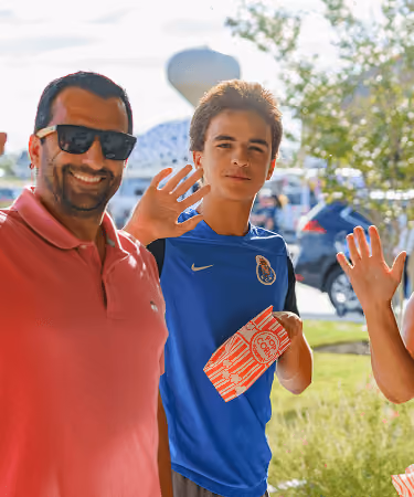 Smiling man in sunglasses and boy in blue shirt waving outdoors on a sunny day.