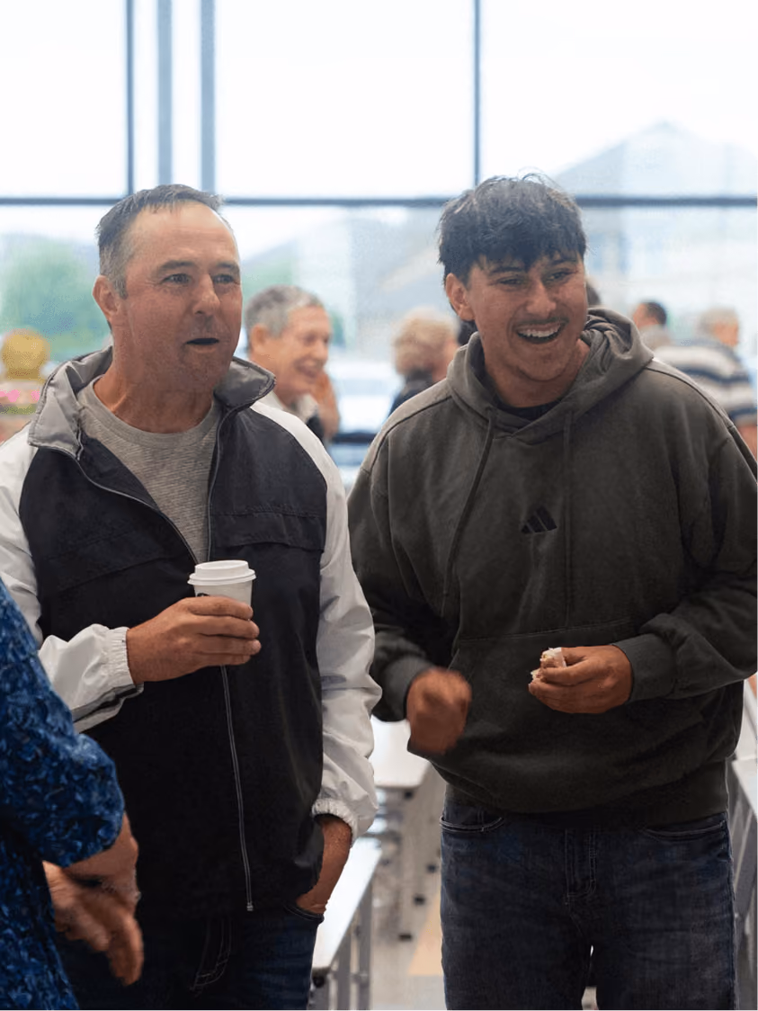 Two men standing indoors, one holding a coffee cup, both smiling and engaging in conversation.