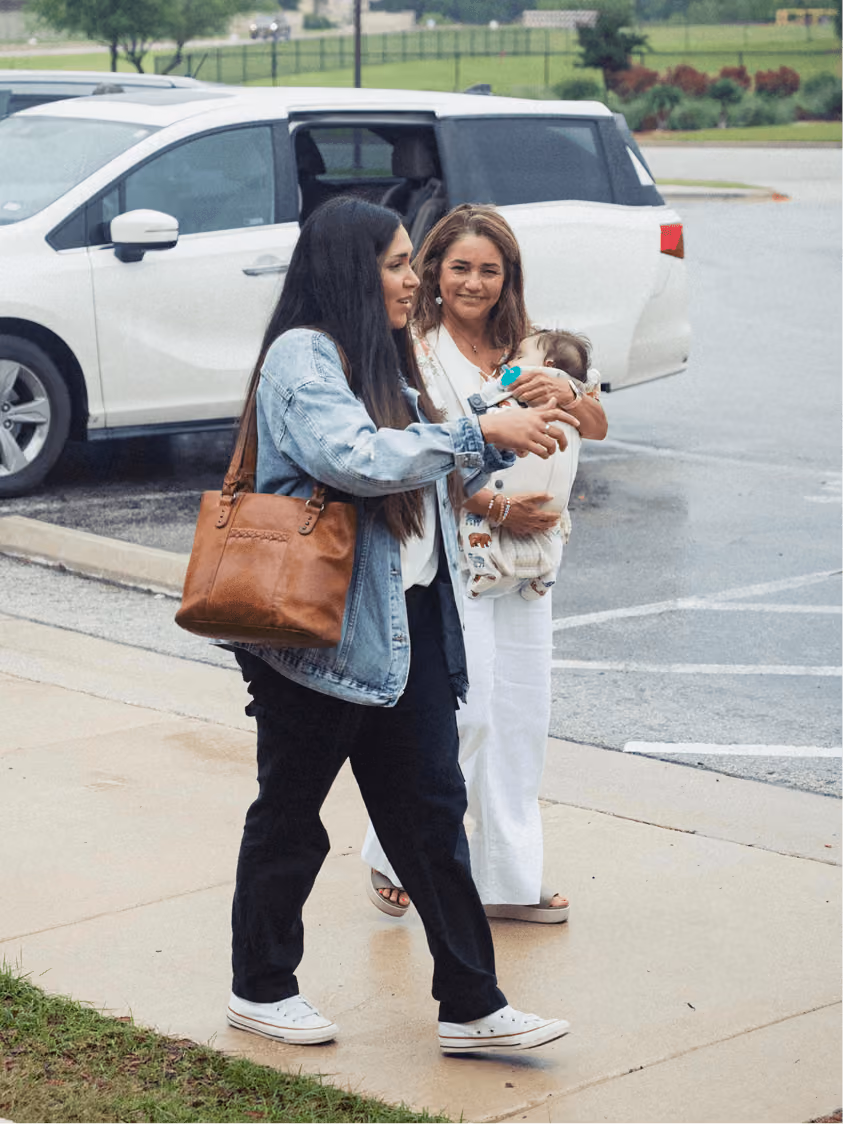 Two women walking on sidewalk near white car; one woman carries a baby with a pacifier.