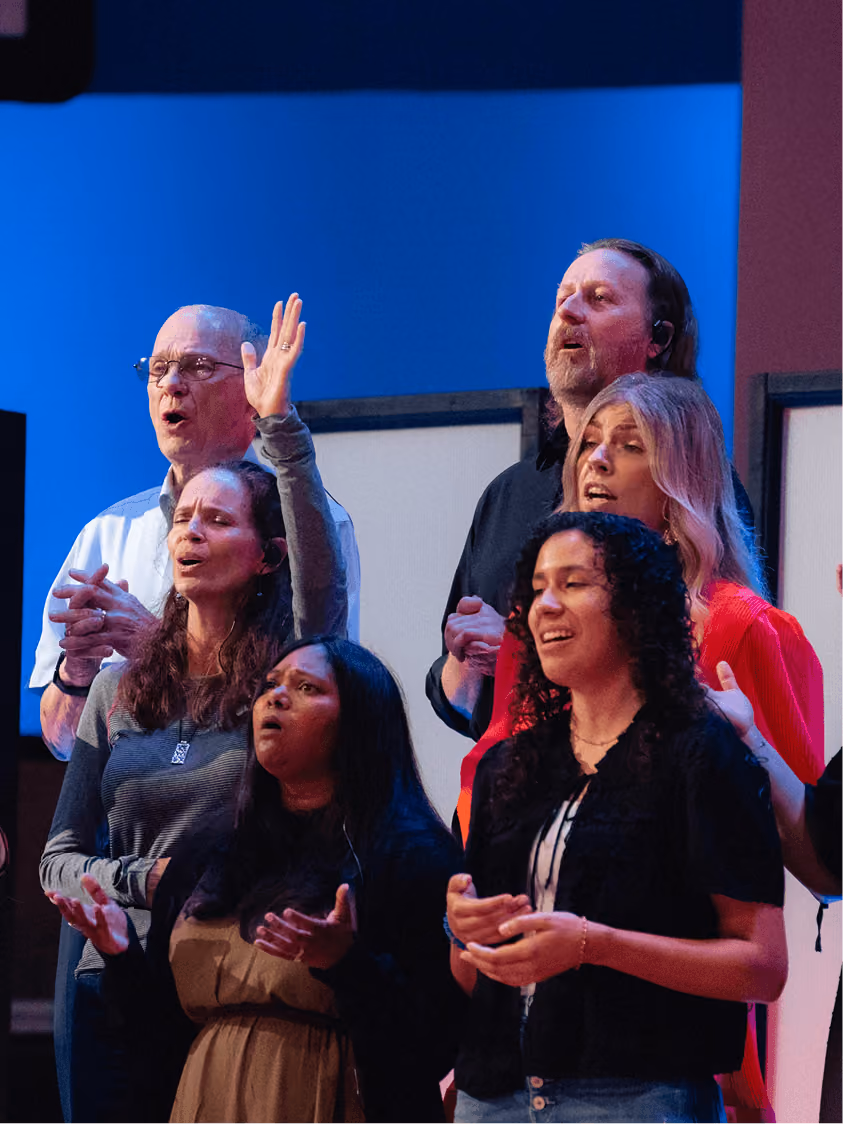 Group of six people singing with expressive faces and raised hands on stage with blue background.
