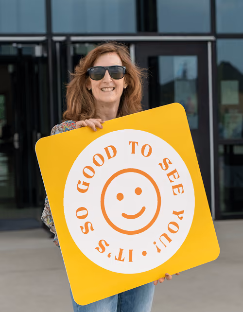 Smiling woman with sunglasses holding a yellow sign reading It's so good to see you with smiley.