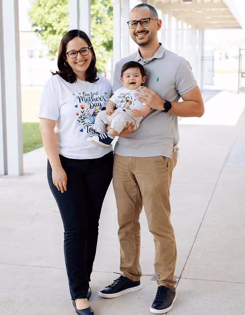 Smiling family of three outdoors; mother wears a Mother's Day shirt holding a baby, father stands beside.