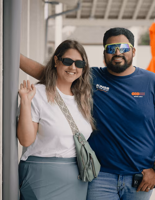 Smiling man and woman wearing sunglasses standing close together, woman waving with handbag over shoulder.