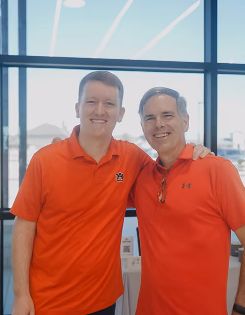 Two smiling men in orange shirts standing arm in arm inside a building with large windows.