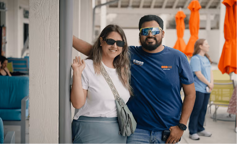 Smiling man and woman wearing sunglasses standing close together outdoors near orange umbrellas.