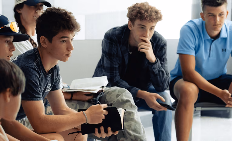 A group of teenage boys sitting and reading books, some holding open Bibles during a discussion.