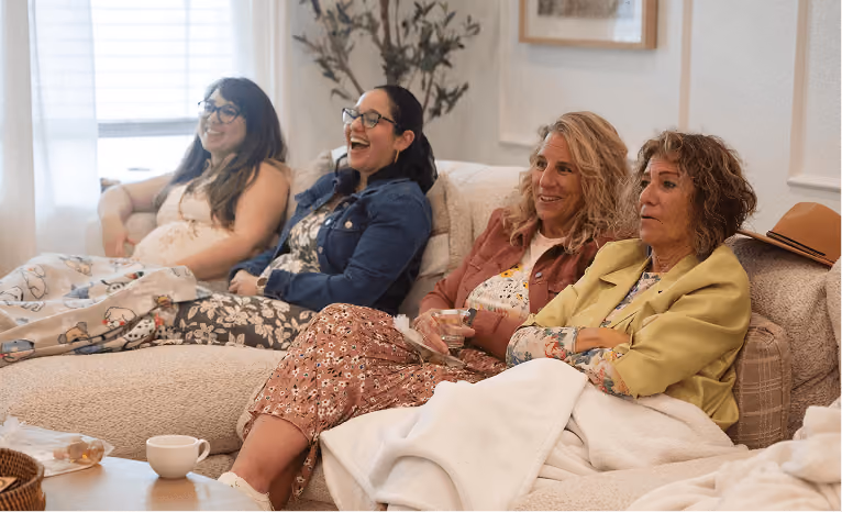 Four women sitting together on a couch, smiling and engaged in conversation in a cozy living room.