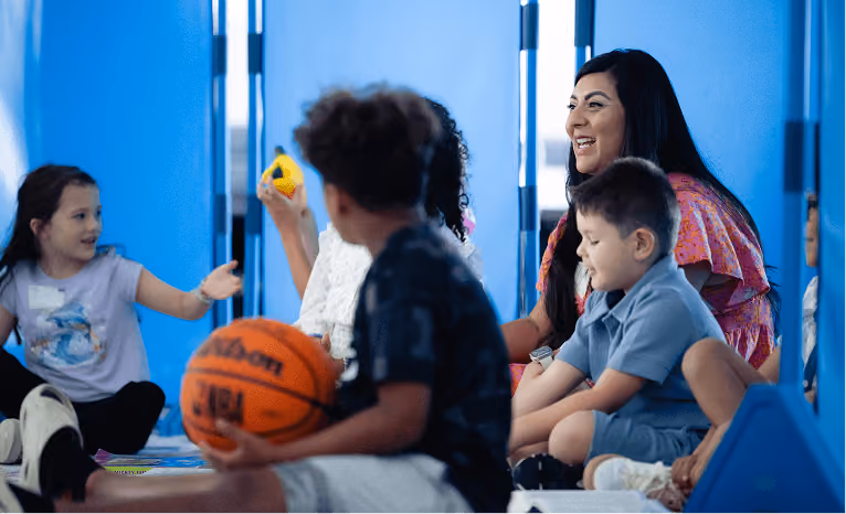 A woman and diverse children sit and engage in a playful group activity in a bright blue room.