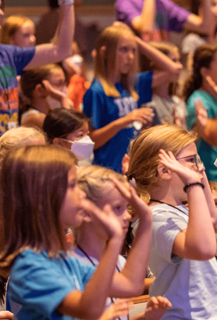Group of children in colorful shirts raising hands indoors, one child wearing a face mask.