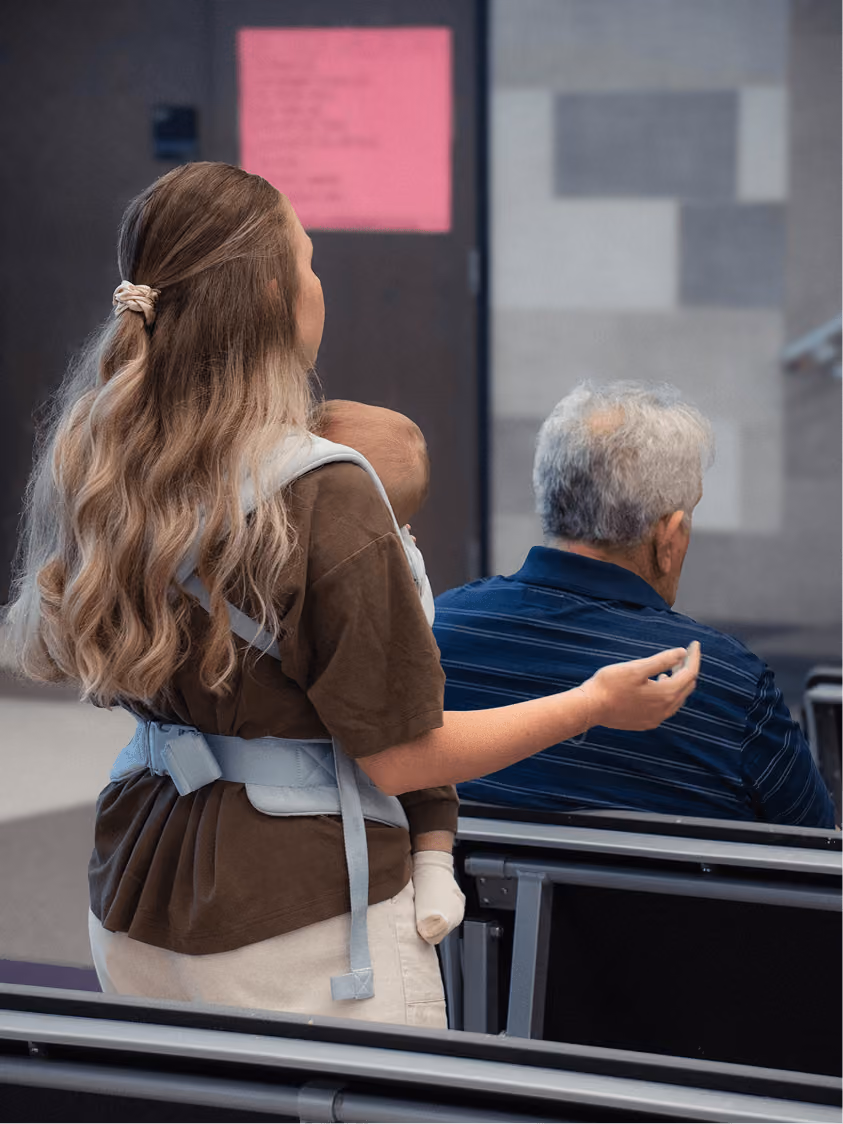 Woman holding a baby in a carrier gestures while sitting next to an elderly man in a waiting area.