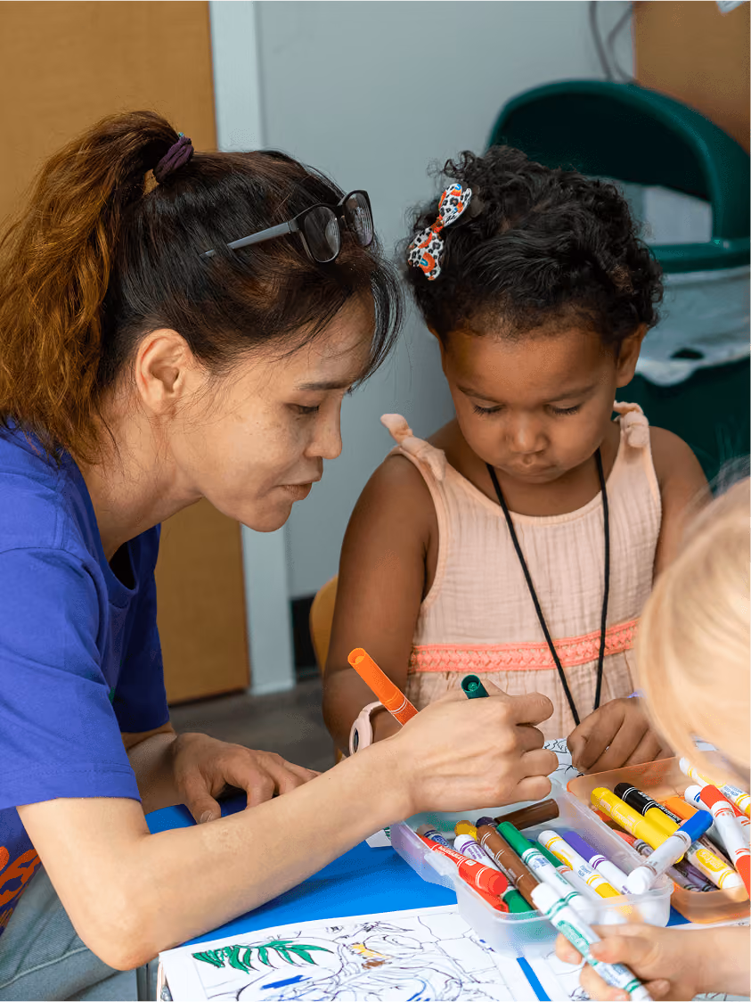 Woman helping young child color at a table with markers and coloring sheets.
