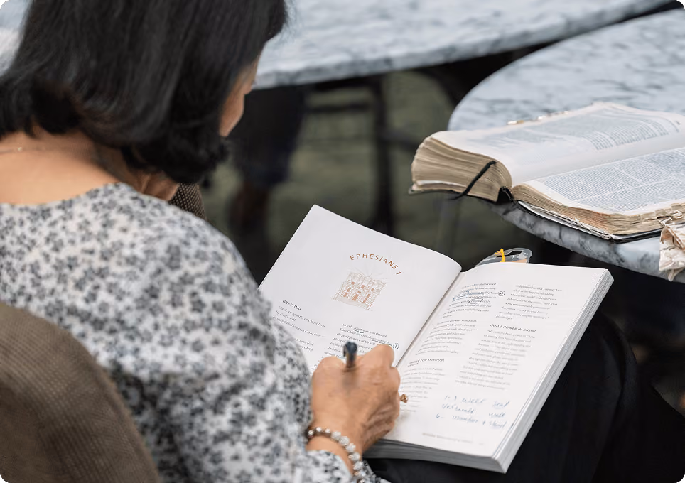 Person writing notes in an open book titled 'Ephesians 1' with another open book on a marble table nearby.