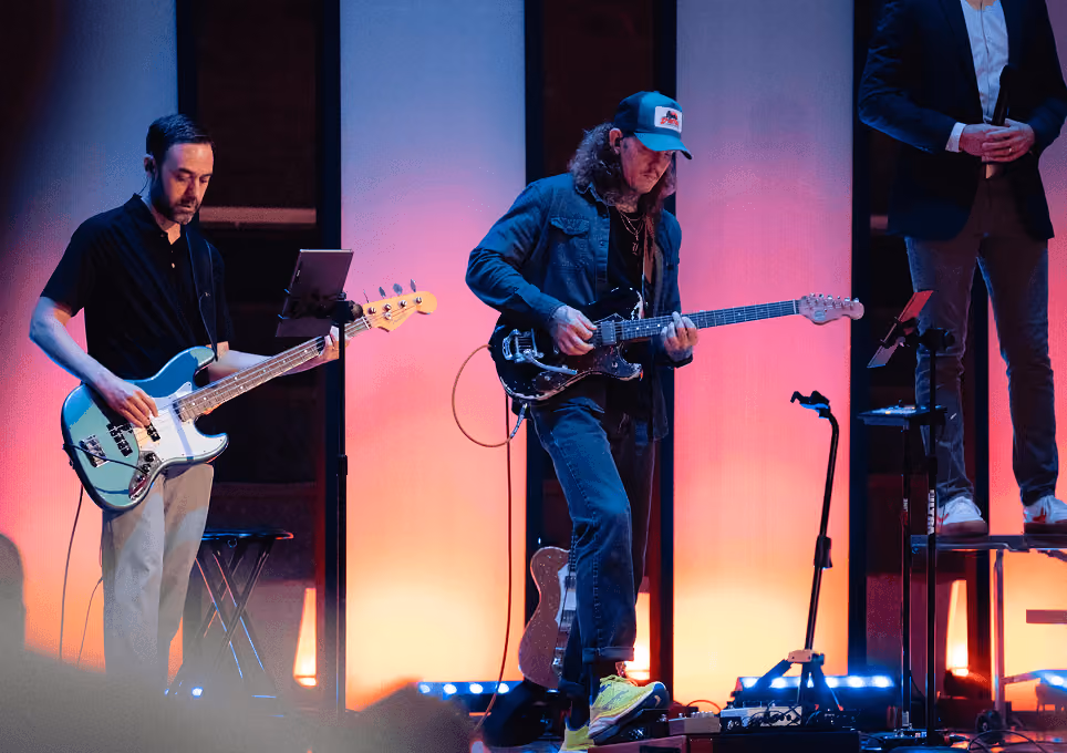 Two musicians playing electric guitars on stage with colorful vertical light panels behind them.