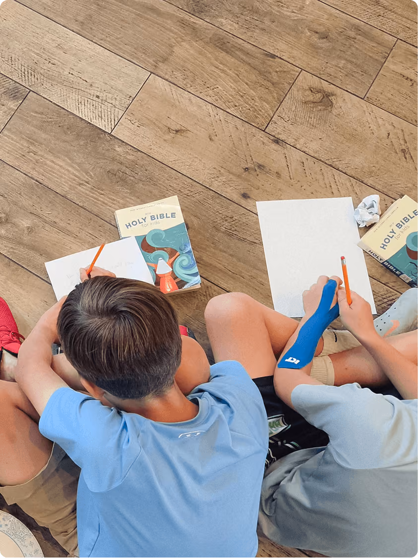 Two children sitting on wooden floor writing with pencils next to 'Holy Bible for Kids' books.