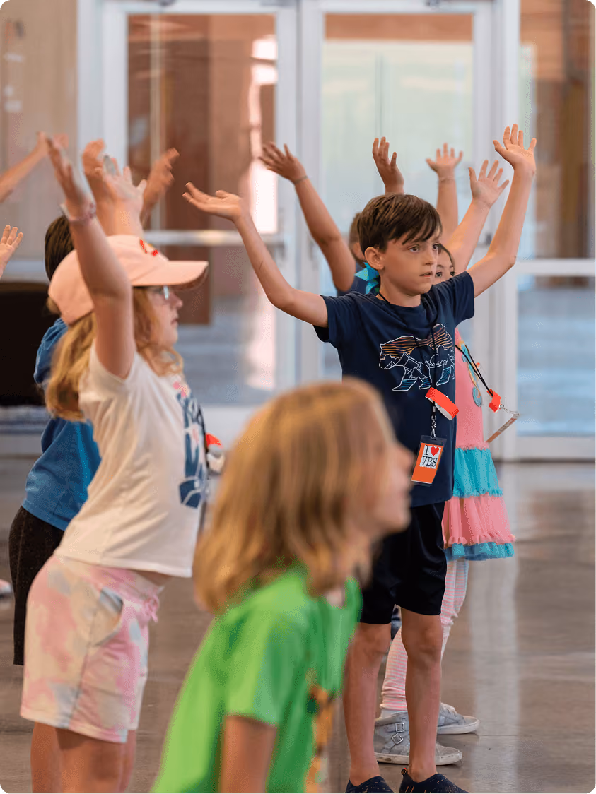 Children standing with arms raised indoors, participating in an activity or exercise.