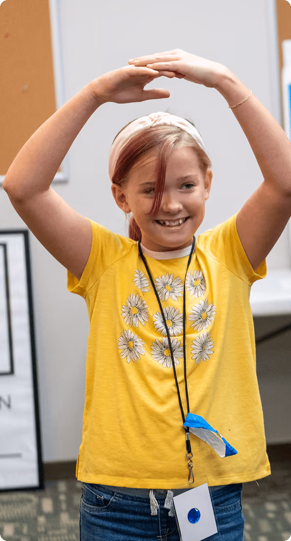 Smiling child in yellow daisy shirt raising arms above head in a curved shape indoors.