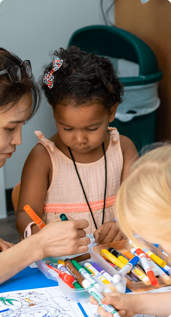 A young girl with a bow in her hair concentrates on coloring with markers guided by an adult.