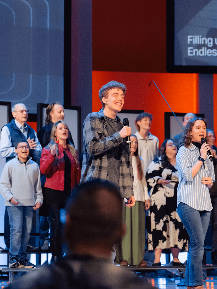 A group of people singing on stage, two holding microphones, with an audience member in foreground.