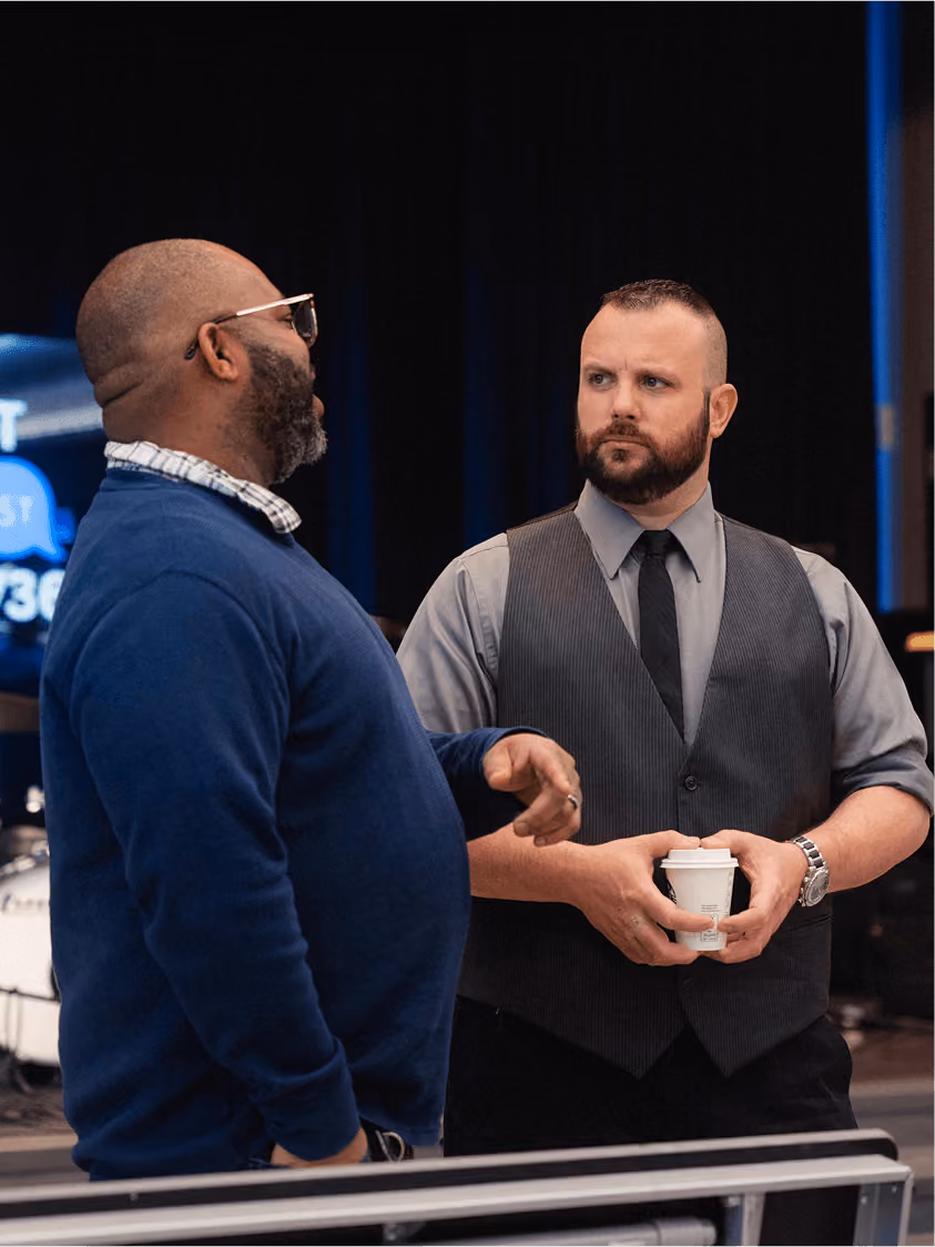 Two men engaged in conversation, one in a blue sweater, the other in a gray shirt and vest holding a cup.