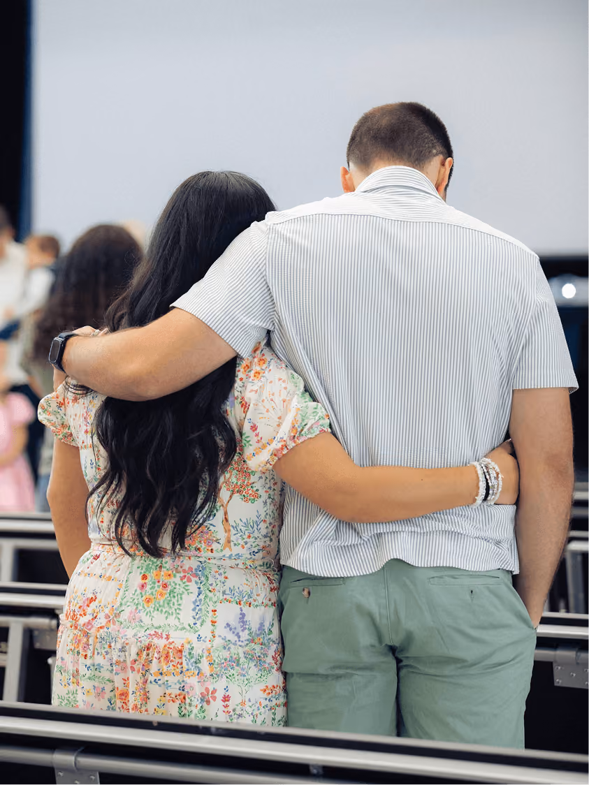 Couple standing with backs to camera, arms around each other in a casual indoor setting.