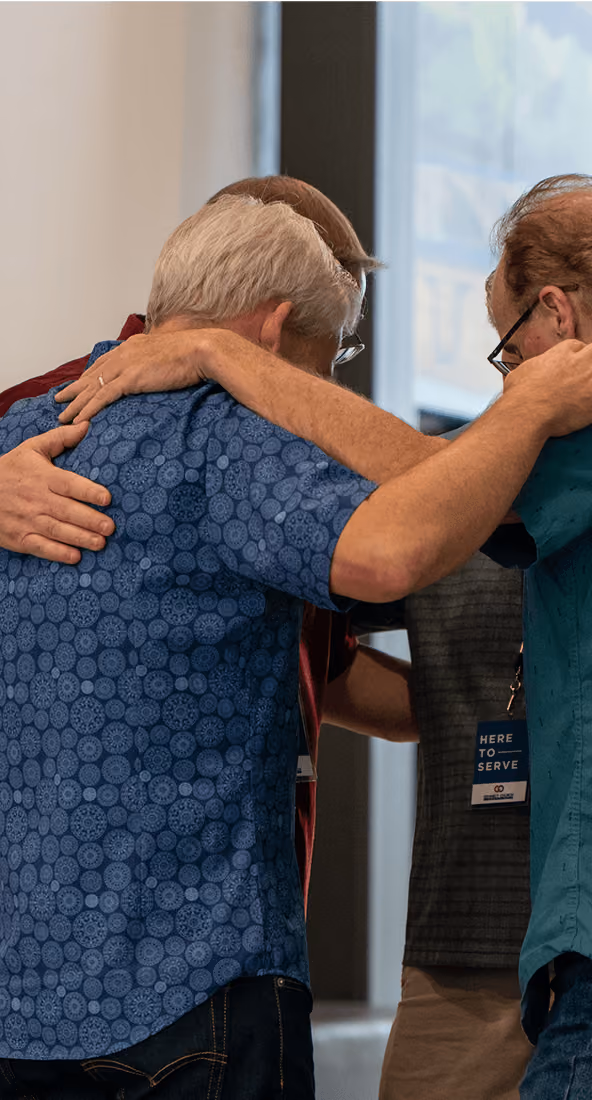 Three men in a group hug with heads bowed in an indoor setting.