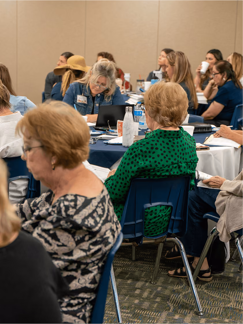 Group of adults seated around tables in a classroom or seminar setting, engaged in writing and reading.