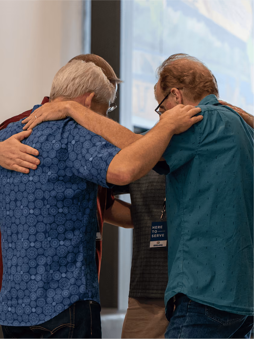Three older men standing close with arms around each other in a group hug.