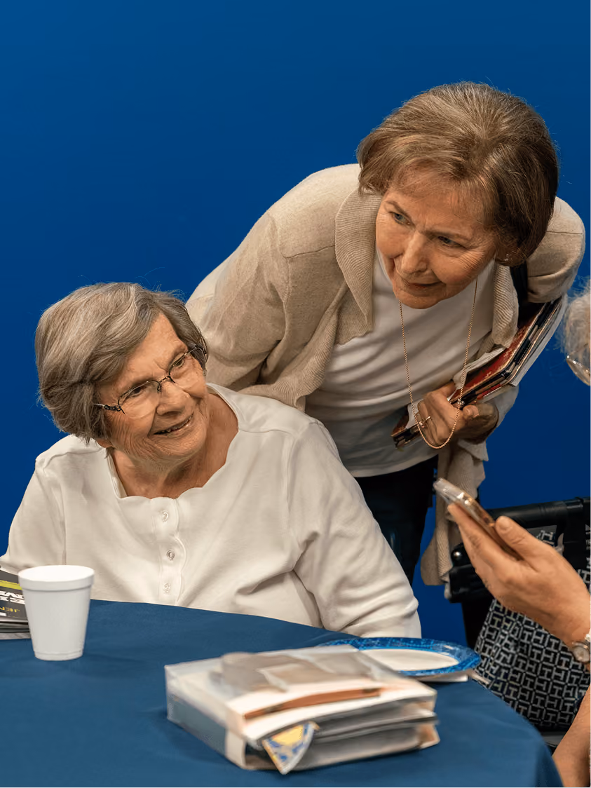 Two elderly women smiling and talking at a table with folders, cup, and smartphone visible.