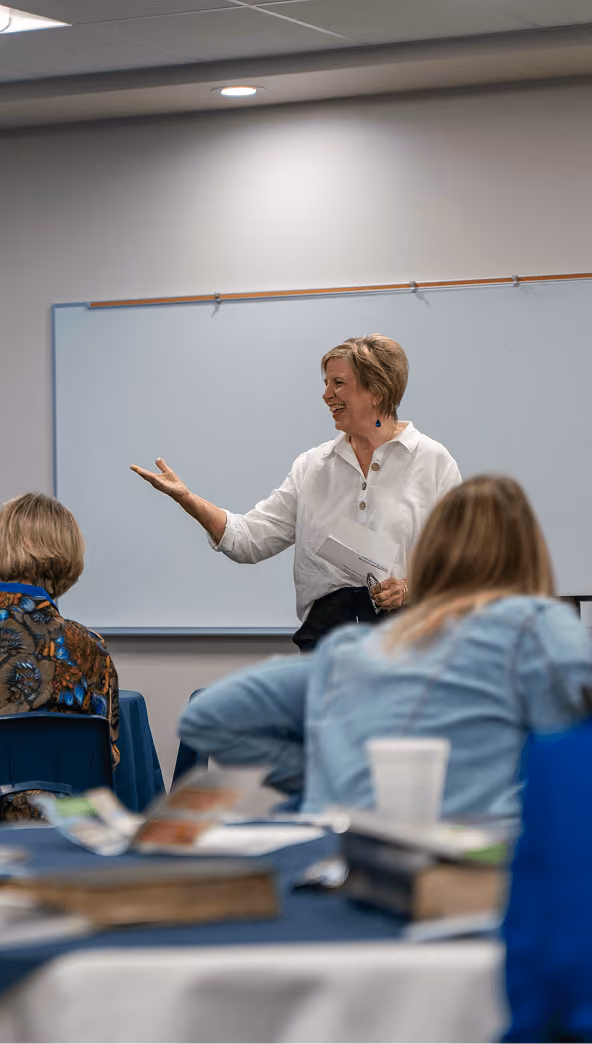 Woman speaking and gesturing in front of a whiteboard to an adult classroom.