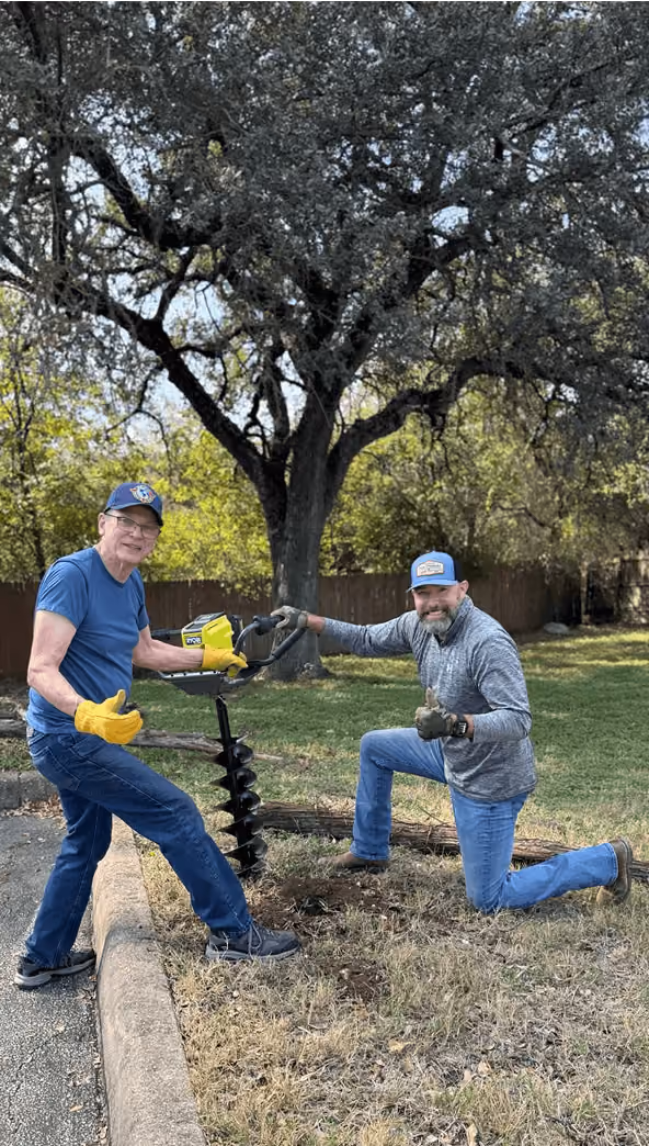 Two men outdoors using a yellow earth auger to drill a hole in the ground near a tree.