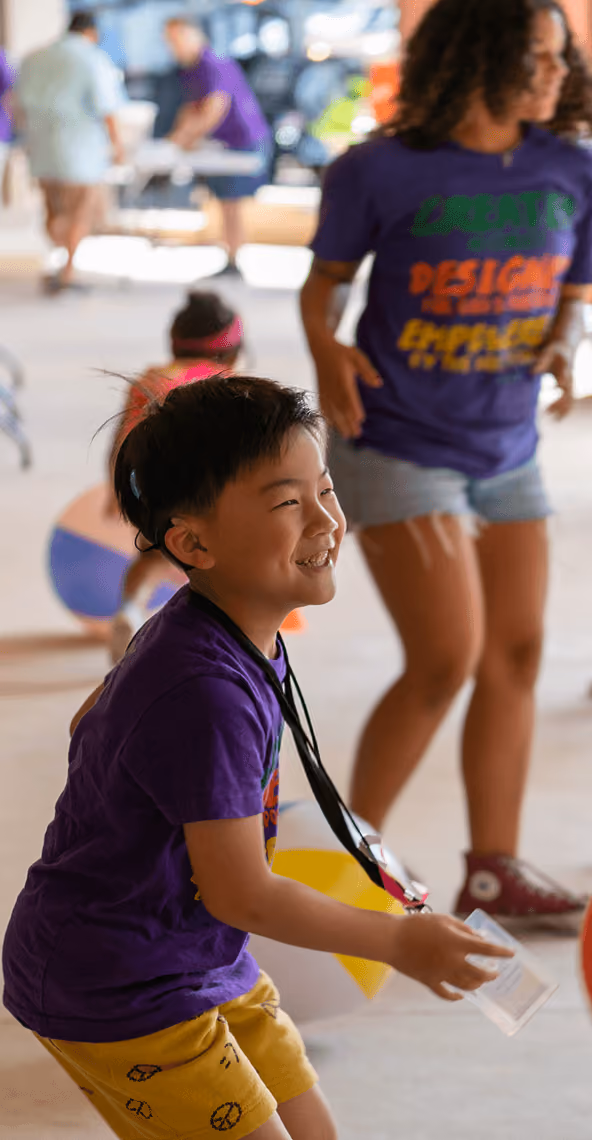 Smiling boy in purple shirt and yellow shorts playing at an indoor event with other children.