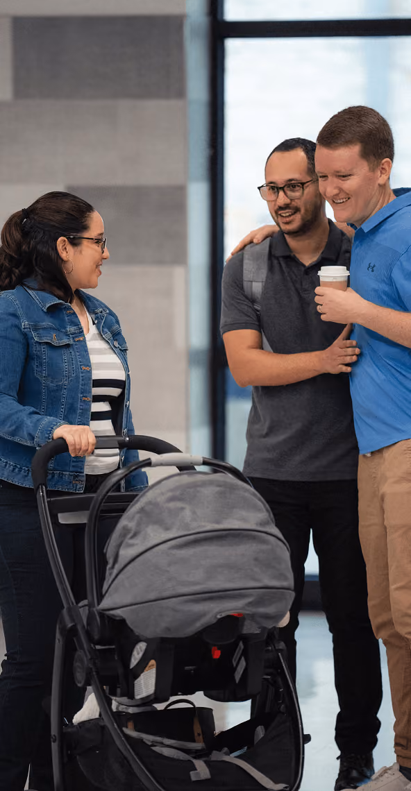Three adults smiling and talking, one pushing a baby stroller, indoors near a window.