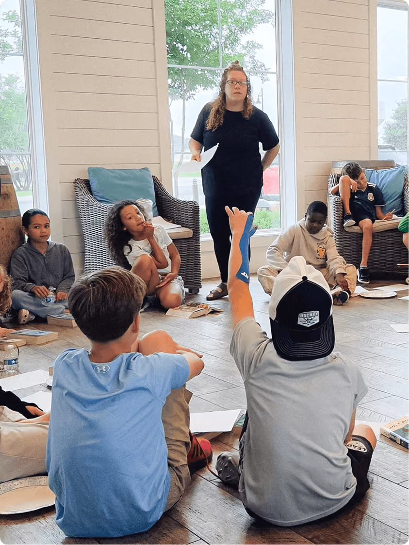 Teacher standing with paper while children sit on floor and chairs, one child raising hand.