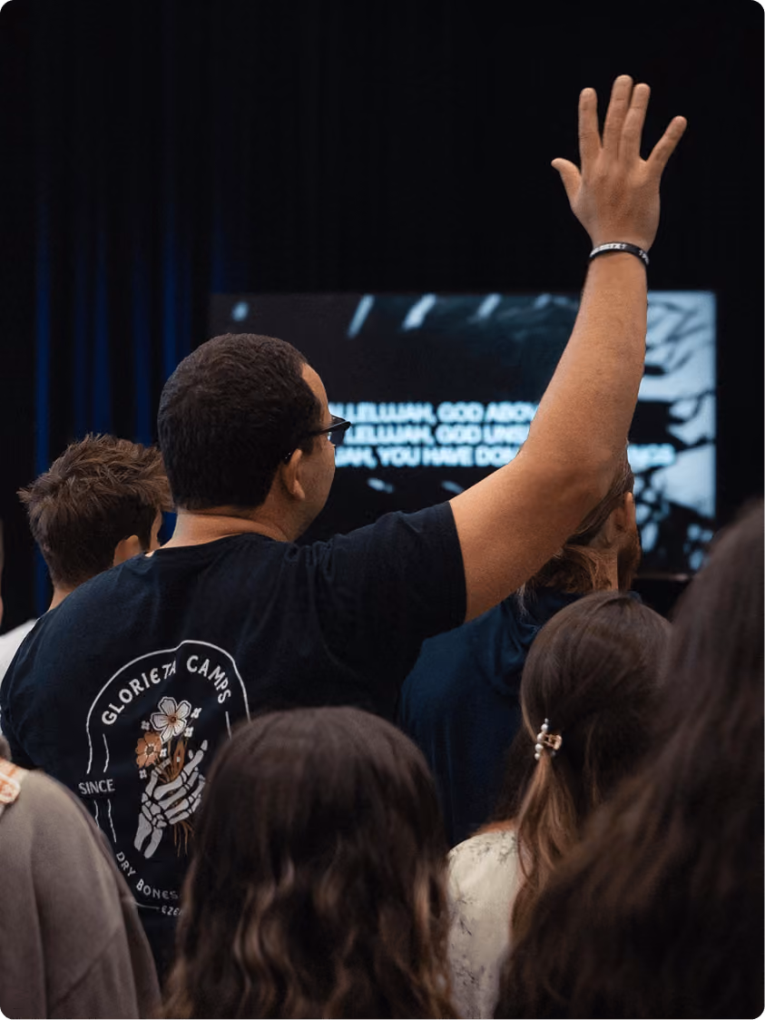 Man wearing glasses and a Glorieta Camps shirt raises his hand among a crowd at an event.