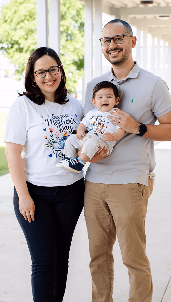Smiling family of three, mother and baby in matching Mother's Day shirts, father holding the baby outdoors.