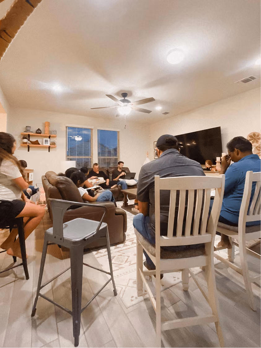 Group of people seated in a living room with a ceiling fan and large TV on the wall.