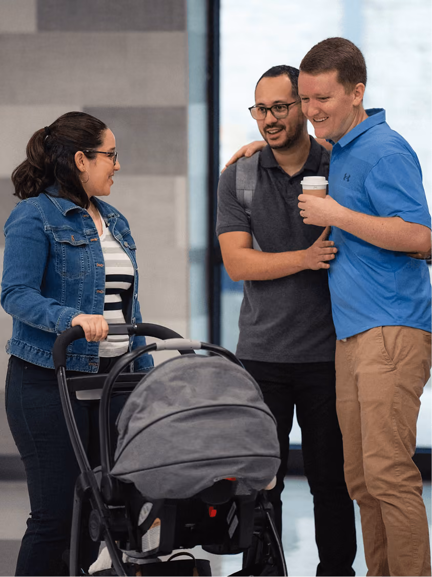 Three adults smiling and talking, one pushing a baby stroller, one holding a coffee cup indoors.