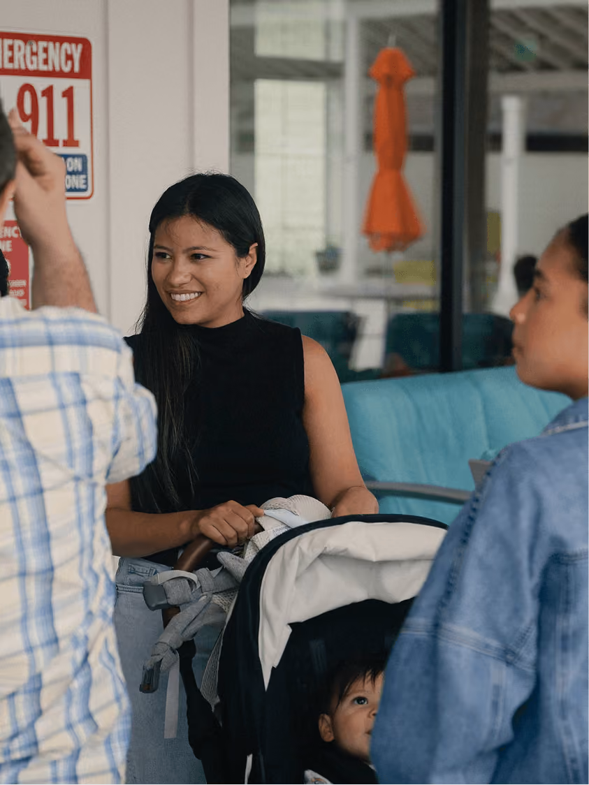 Smiling woman with long dark hair standing behind a baby stroller, talking to two people nearby.