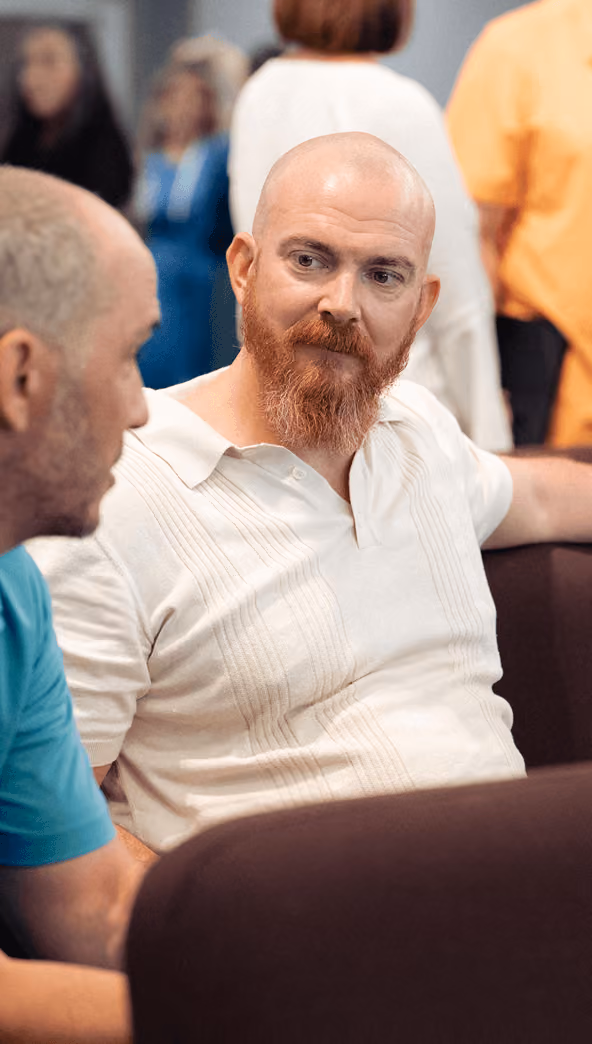 Bald man with red beard in white shirt attentively talking to another man in a blue shirt indoors.