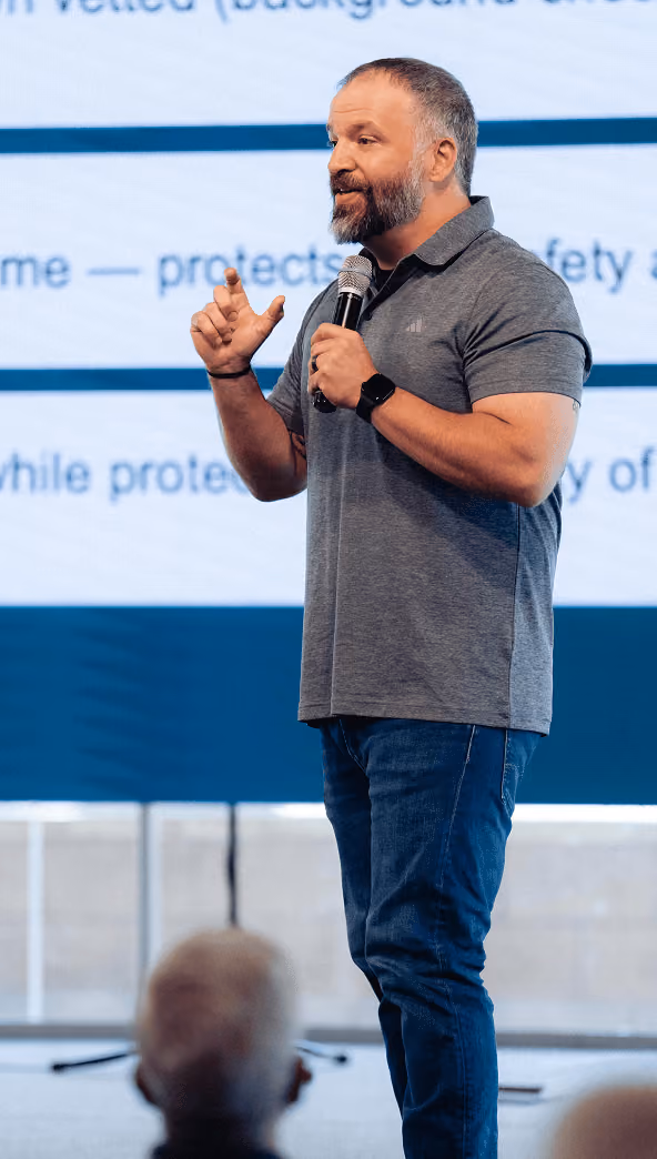 Man with beard holding microphone and speaking on stage with blurred audience in foreground.