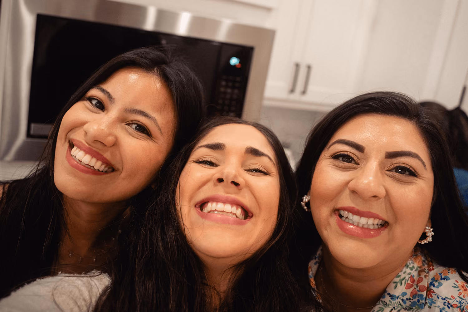 Three smiling women close together taking a selfie in a kitchen with a microwave behind them.