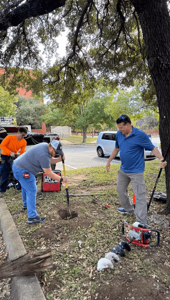 Three men dig a hole near a tree in an outdoor urban area with tools and equipment nearby.