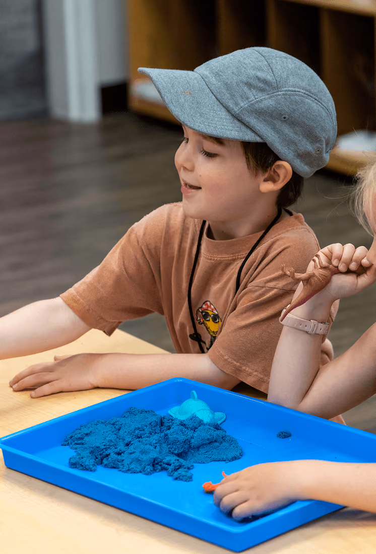 Boy in gray cap playing with blue kinetic sand and toy dinosaurs on a blue tray.