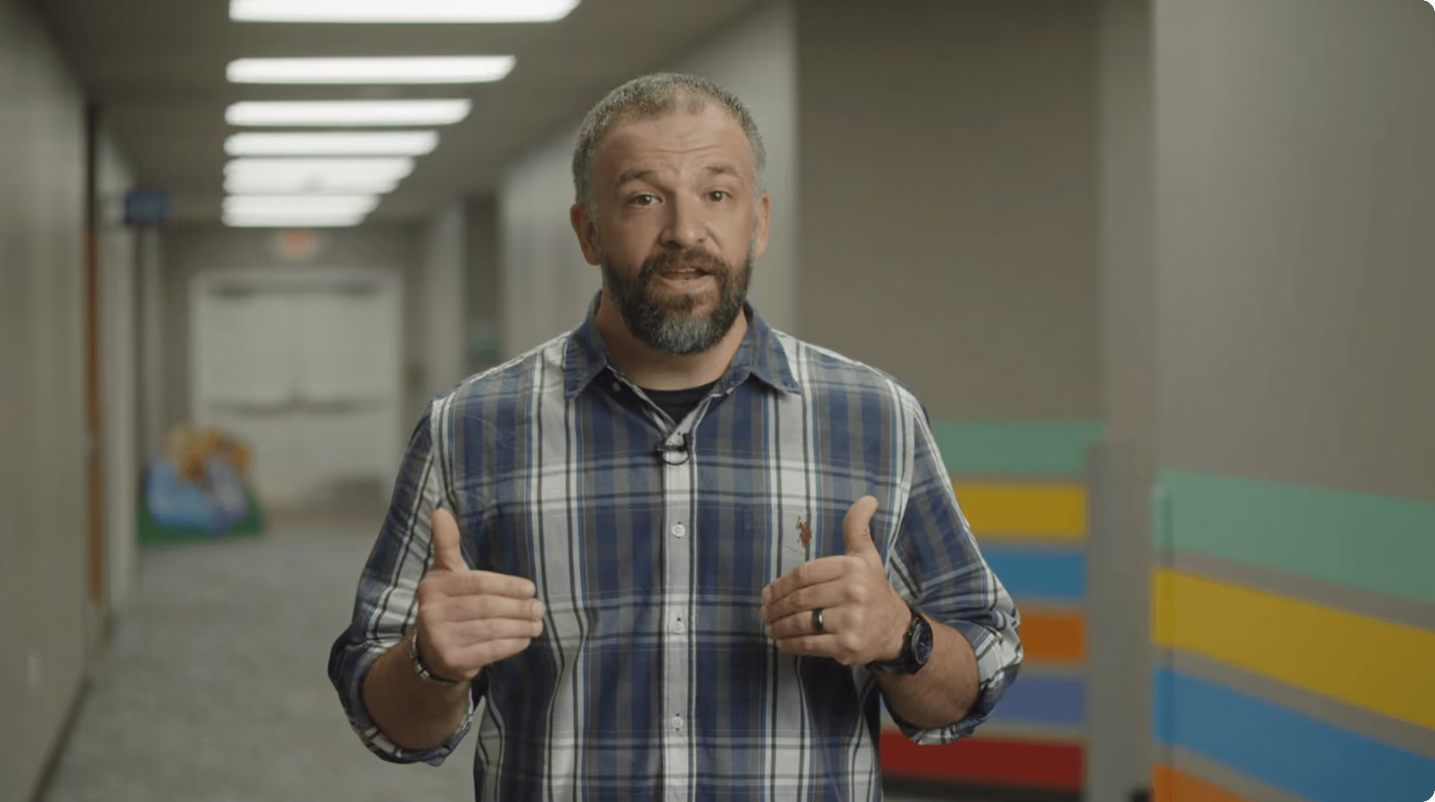 Bearded man in a blue plaid shirt speaking with hand gestures in a hallway with colorful wall stripes.